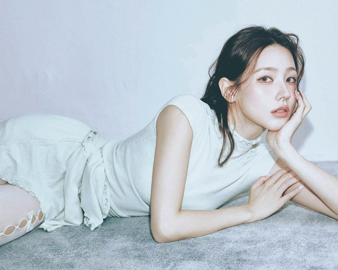 Woman lying on a gray surface with a plain background, wearing a white sleeveless top and light-colored pants.